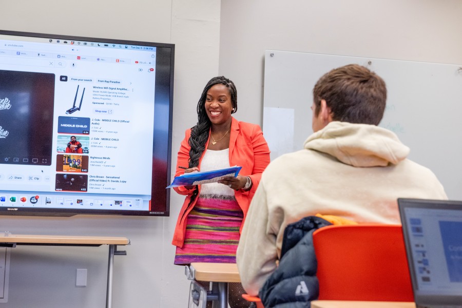 Person giving a speech in front of a class of students with a digital presentation behind them.
