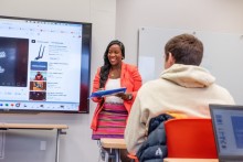Person giving a speech in front of a class of students with a digital presentation behind them.