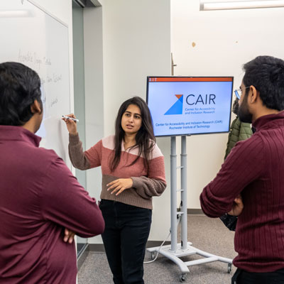 a woman writes on a whiteboard and speaks to two men