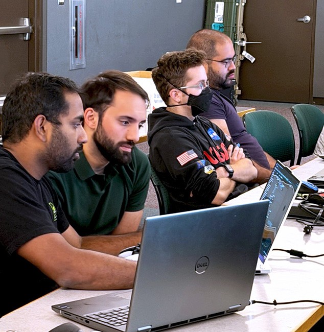Four people sit at a table with laptops in front of them. Two of them are looking at something on a laptop screen while the other two are watching a presenter out of frame.