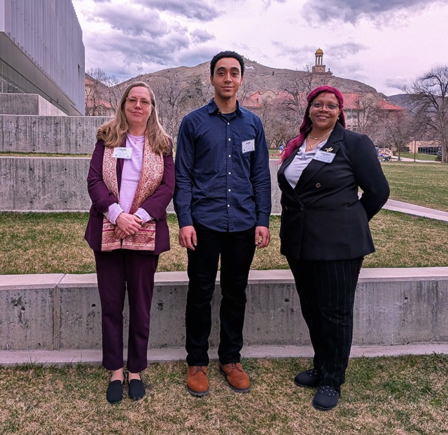 Three people stand in front of tiered concrete and grass terrace. A clock tower and mountain rise in the background.
