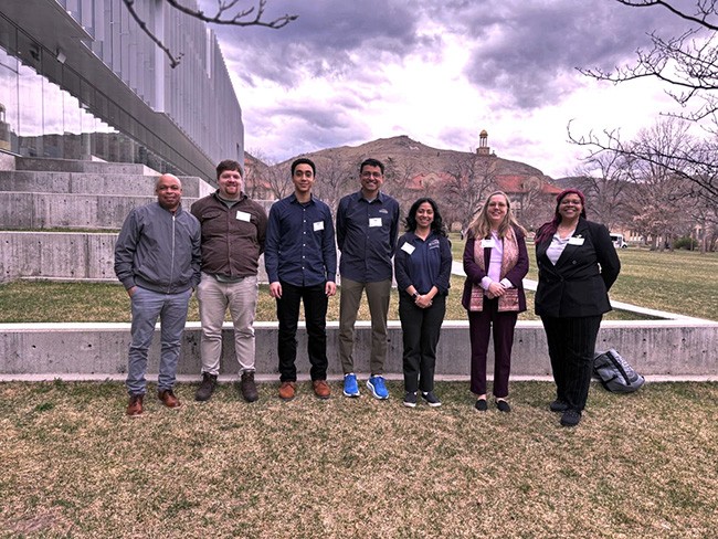 Seven people stand in front of tiered concrete and grass terrace. A clock tower and mountain rise in the background.