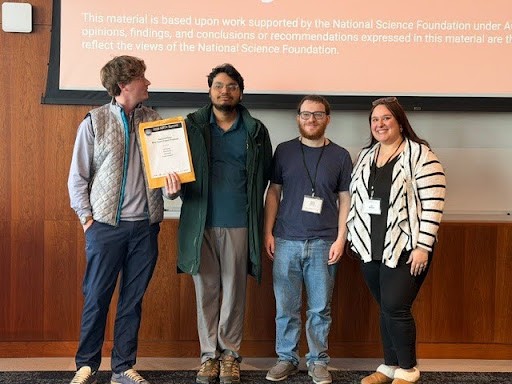 Four people stand on a in front of a projector screen next to a podium, smiling. One person on the left looks towards the projector screen while holding an award certificate.
