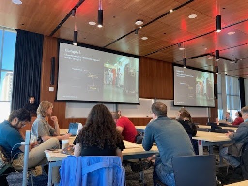 A room full of people sit at tables watching two overhead screens during a presentation.