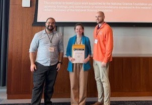 Three people stand on a in front of a projector screen next to a podium, smiling. The person in the center is holding an award certificate.