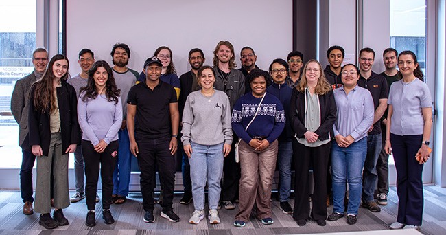 A group of smiling graduate students and faculty pose for a group photo in front of a projection screen. The group is flanked on either side by two floor-to-ceiling glass doors.