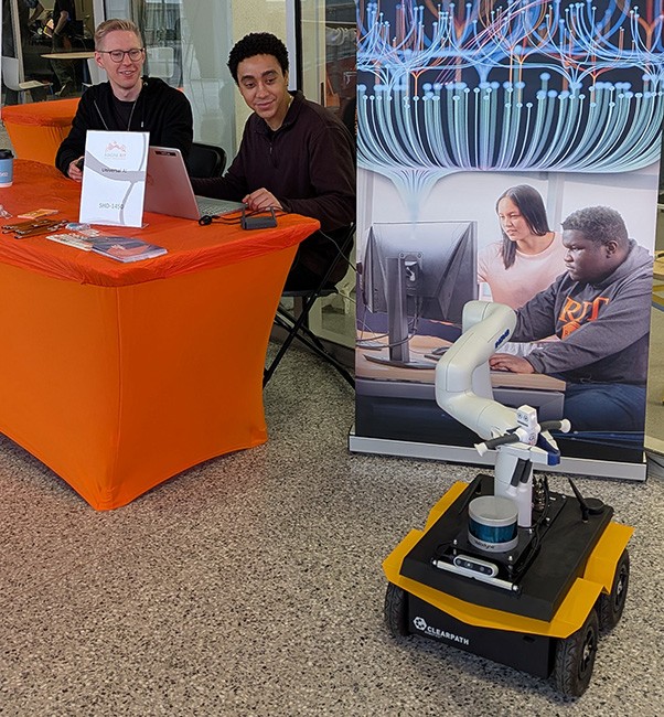 Clearpath Jackal mobile robot with a Kinova robotic arm displayed at a Universal AI exhibit table, featuring information about AI, robotics, and technology research at an RIT technology showcase.