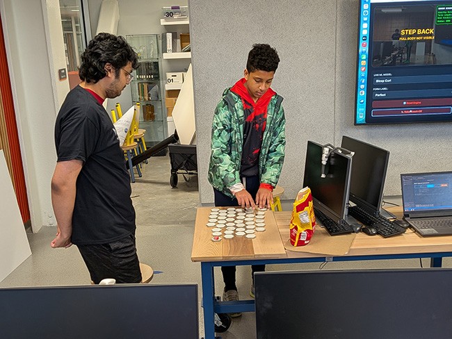 Two people interact with a technology demonstration in a lab, using hexagonal tiles and computer vision software displayed on a large screen.