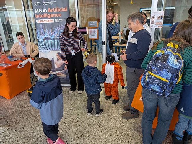 Visitors interact with a small humanoid robot dressed in a tiger costume at an RIT artificial intelligence exhibit featuring robotics and hands-on technology demonstrations