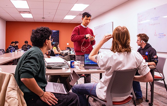 A group of graduate students seated at a table converse with another student standing with them and holding a device with dangling wires. Another group of students are sitting in group in the background