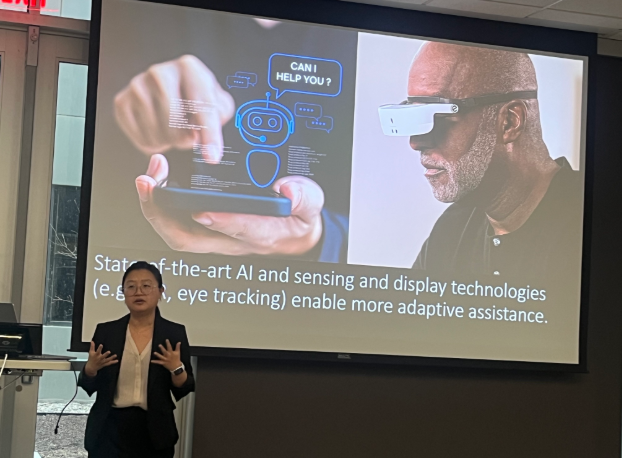 Presenter speaks in front of a large screen displaying AI assistive technology concepts, including a virtual assistant graphic, a person using smart glasses, and text about adaptive assistance through AI sensing and display technologies