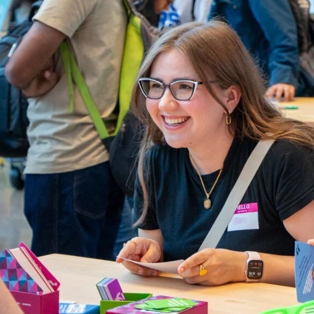seated student holding playing cards, smiles at her teammate during their card game