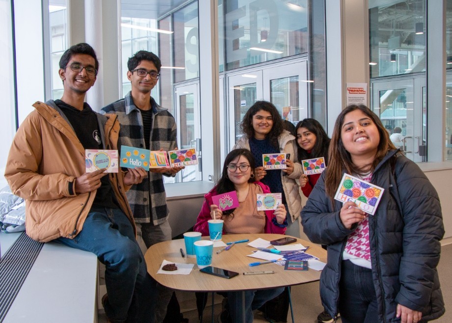 Graduate students sitting at a table holding up postcards they're sending to family smile at the camera