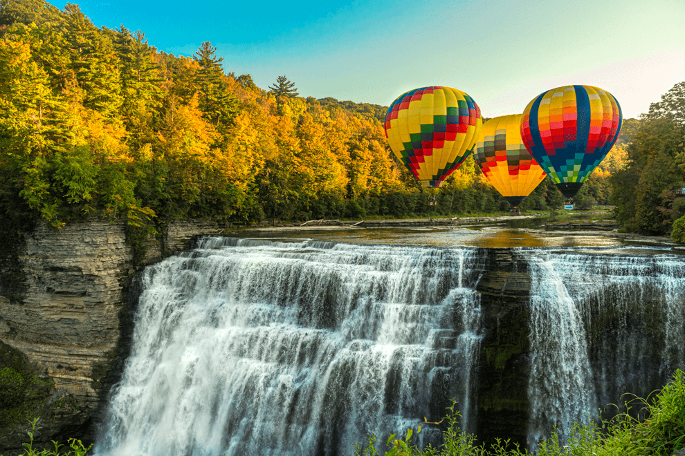 Colorful hot air balloons flying over a large waterfall surrounded by autumn trees.