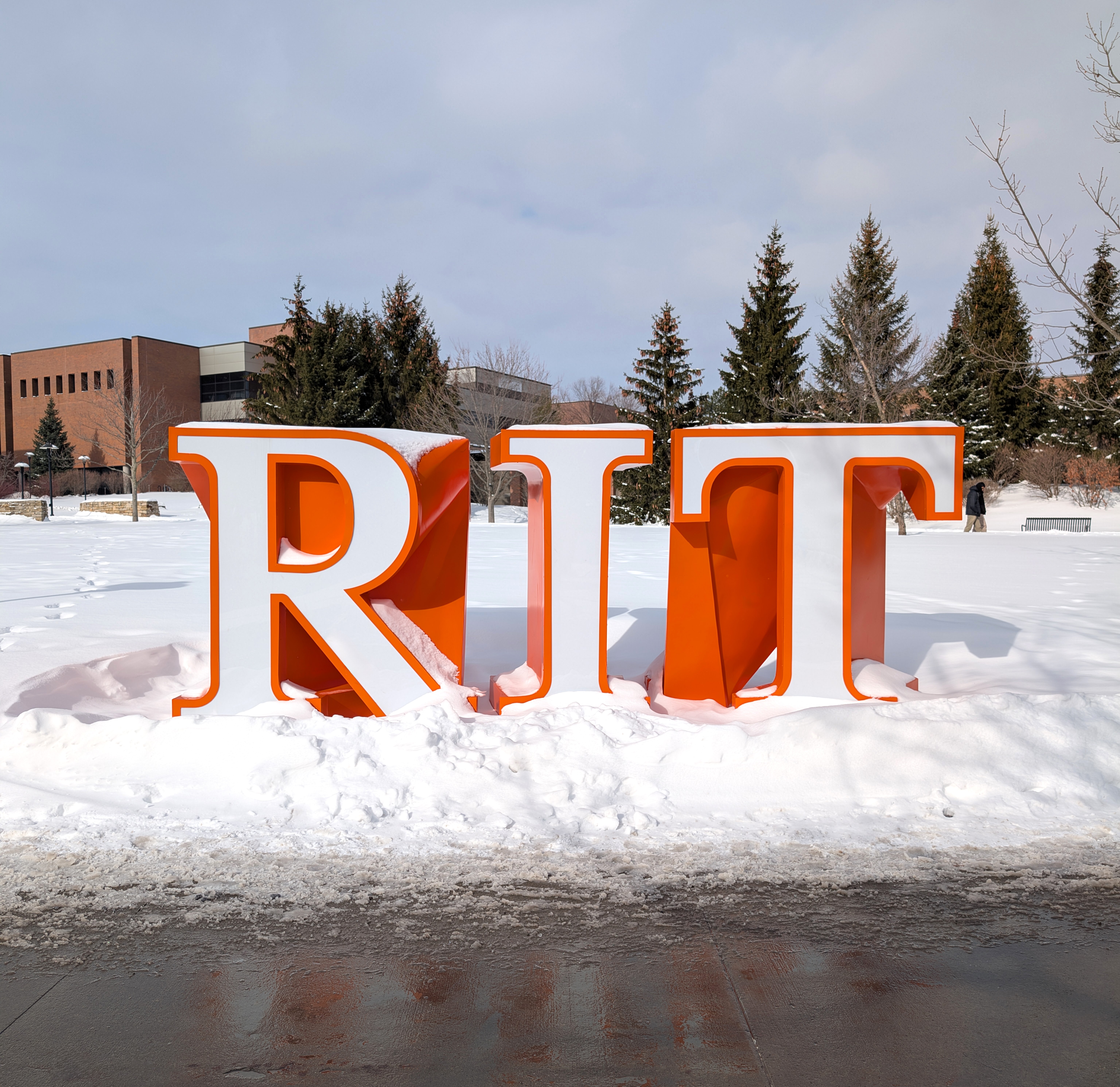 Large orange-and-white “RIT” letters standing in the snow on a winter campus, with trees and brick buildings in the background.