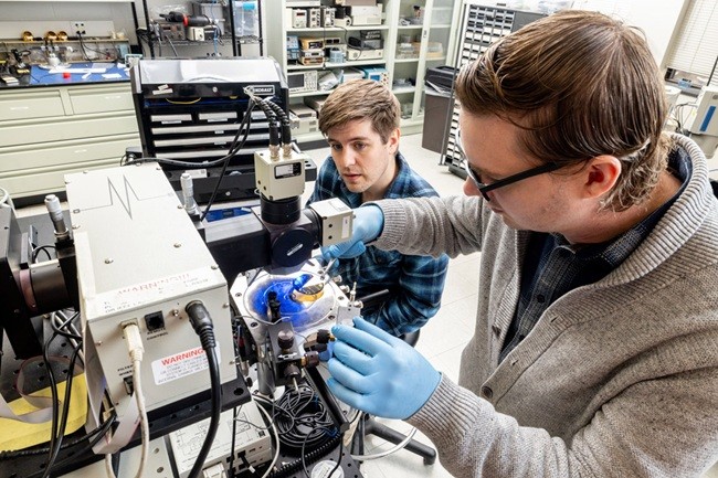Two researchers work together in a laboratory, using gloved hands to adjust scientific equipment and instruments on a lab bench surrounded by tools and electronics.