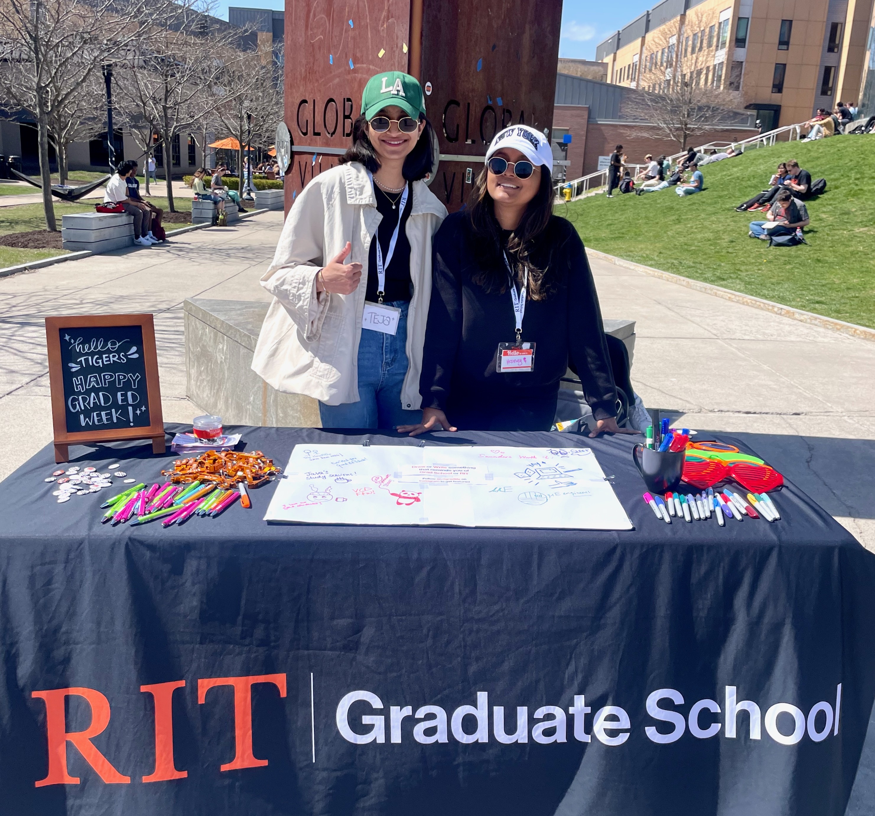 New graduate students sitting on a circular table with one student holding up and showing a gray RIT T-shirt while others smile at the camera