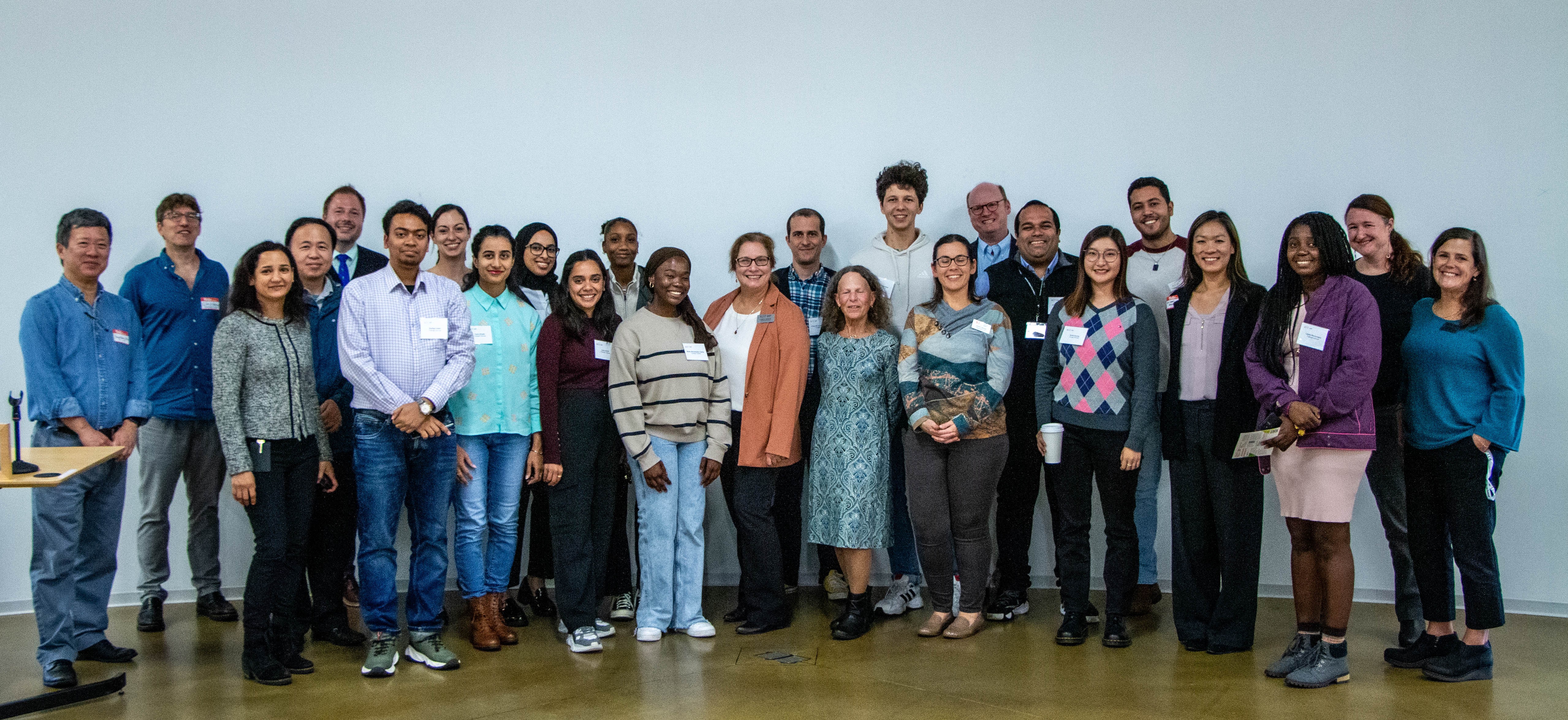 New graduate students sitting on a circular table with one student holding up and showing a gray RIT T-shirt while others smile at the camera