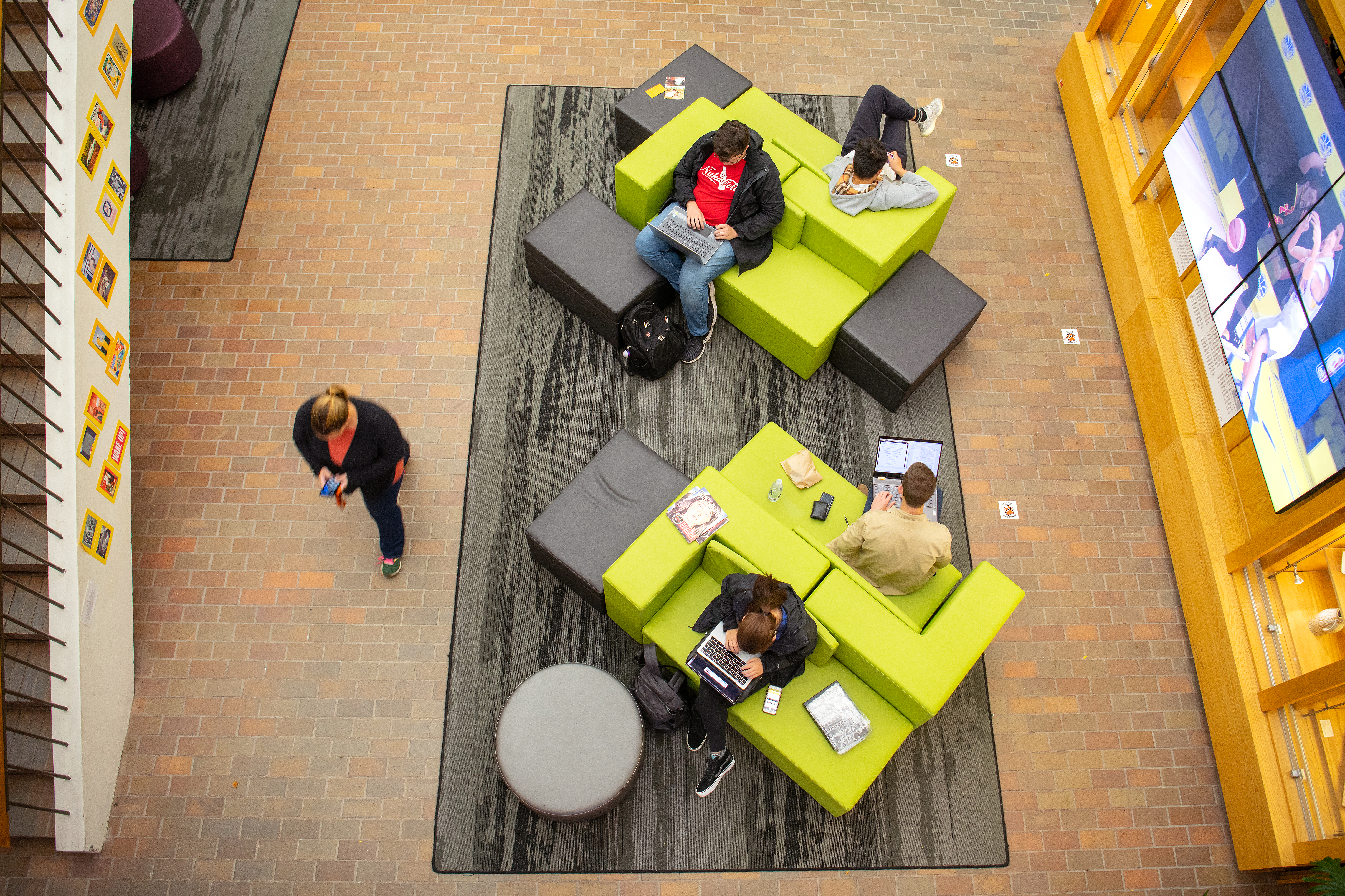 An aerial view shows a common area with brick floors and bright lime green modular sofas. Several students are seated on the sofas, mostly using laptops. A person walks on the left, looking at a phone. To the right, a large digital display is visible.