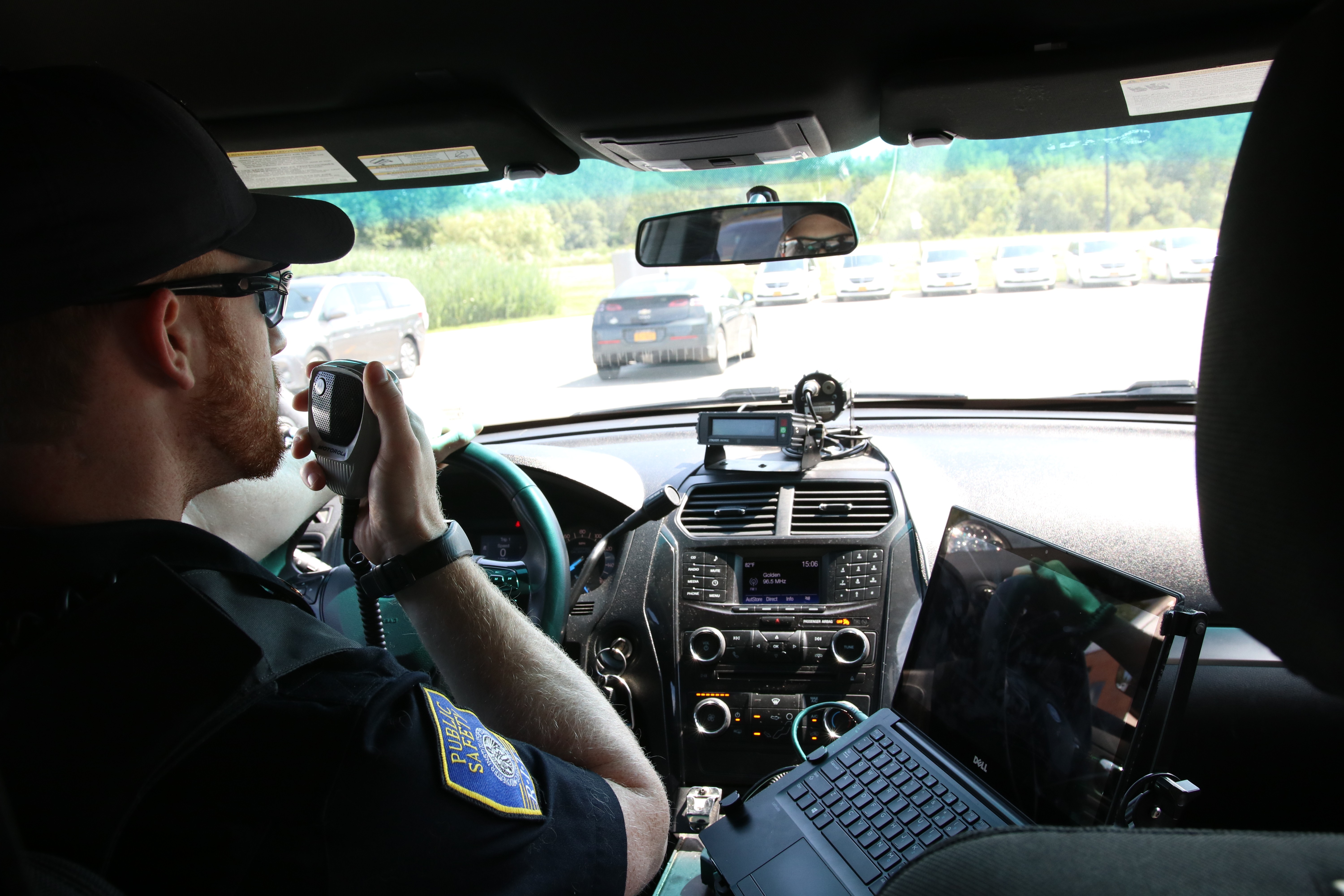 Individual wearing a Public Safety uniform and a black hat his pictured seating in a patrol vehicle using his radio