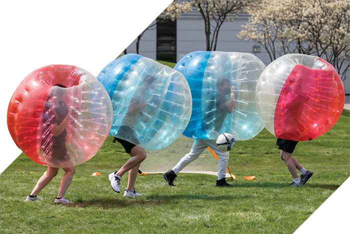 Students run across a grassy field wearing colorful inflatable bubble suits while playing a game.