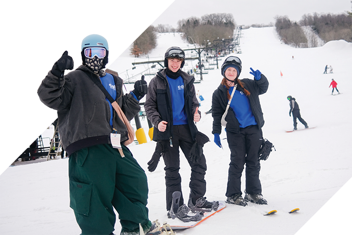 Three students in helmets and winter gear pose with snowboards at the base of a snowy ski slope.