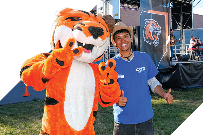 RIT tiger mascot stands beside a student wearing a blue shirt and cowboy hat, giving thumbs up near an outdoor stage setup.