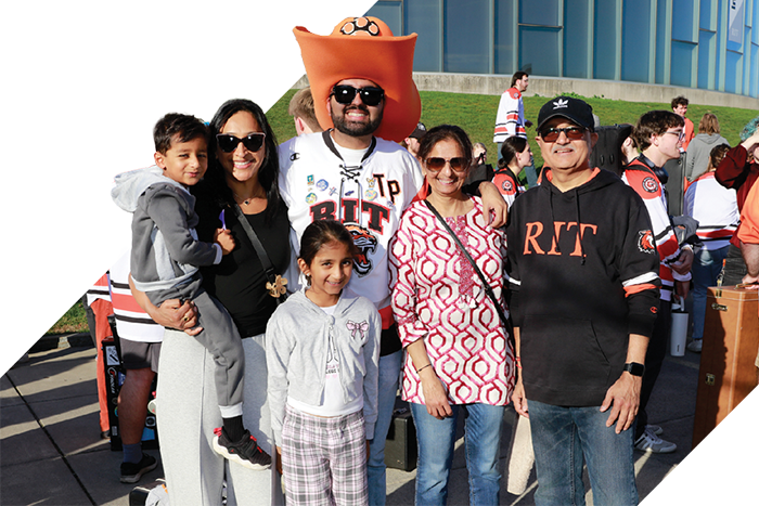 Family group poses outdoors, including a child and an RIT fan wearing a large orange hat, surrounded by others in RIT apparel.