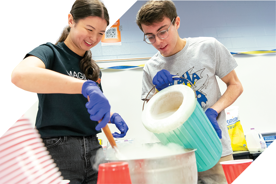 Two students wearing gloves mix ingredients in a large steaming pot, with one pouring from a green container while the other stirs; red cups and supplies sit on the table nearby.