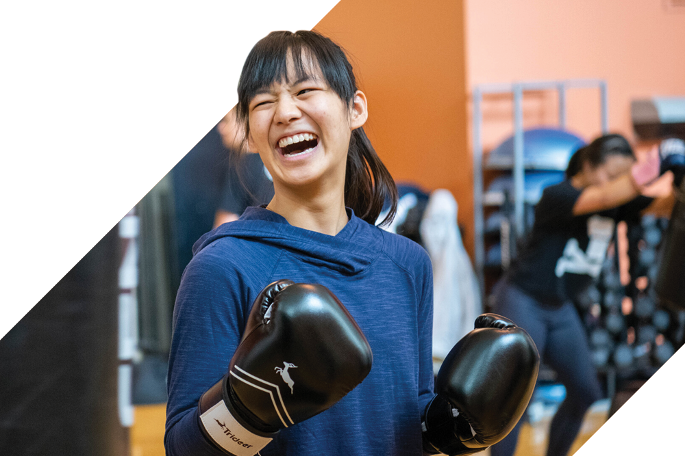 Person in a blue hoodie wearing boxing gloves in a fitness center, with workout equipment and another person training in the background.