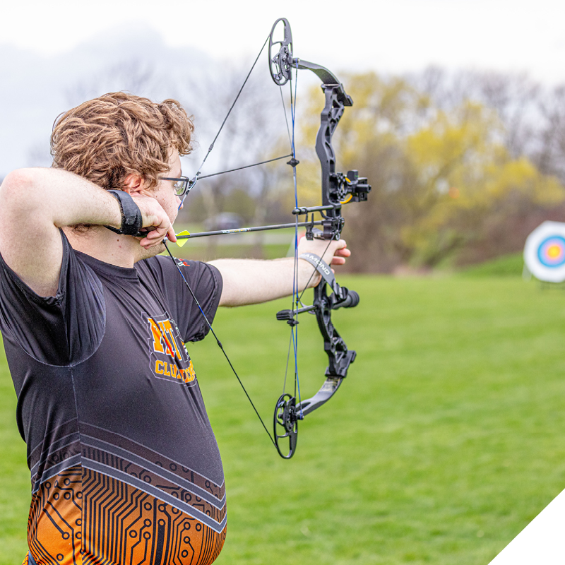 Person aiming a bow and arrow at a target on an outdoor field.