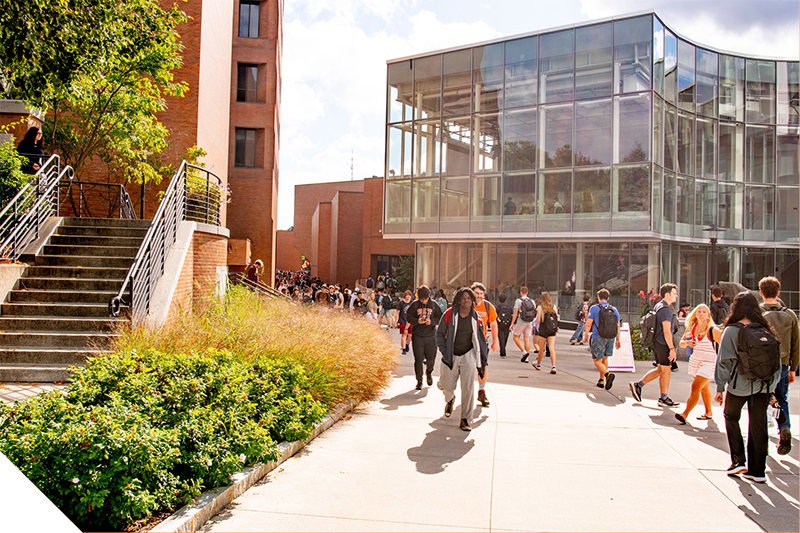 A busy campus walkway outside a modern glass building, with students walking in multiple directions along a wide path. Nearby brick buildings, steps, and landscaping frame the scene on a sunny day.
