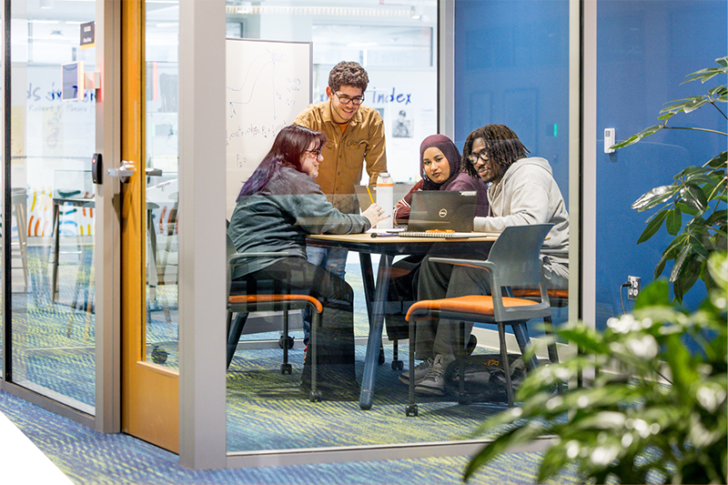 Students work together around a table in a glass-walled study room, looking at a laptop and notes. A whiteboard with writing is visible behind them, and the space includes modern chairs, carpeted flooring, and nearby greenery outside the room.