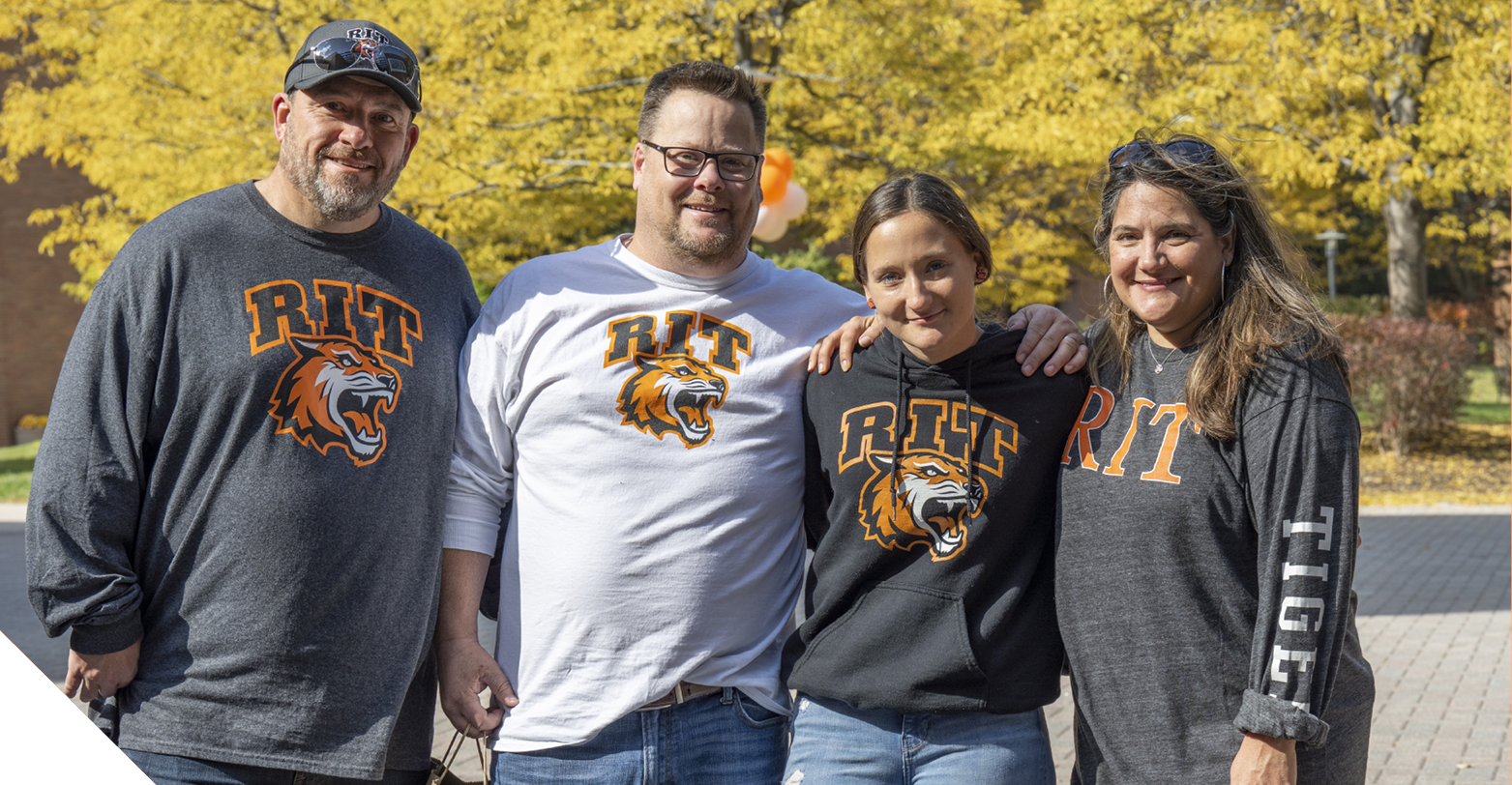 Four people wearing RIT apparel stand together outdoors on campus, with bright yellow fall foliage in the background.