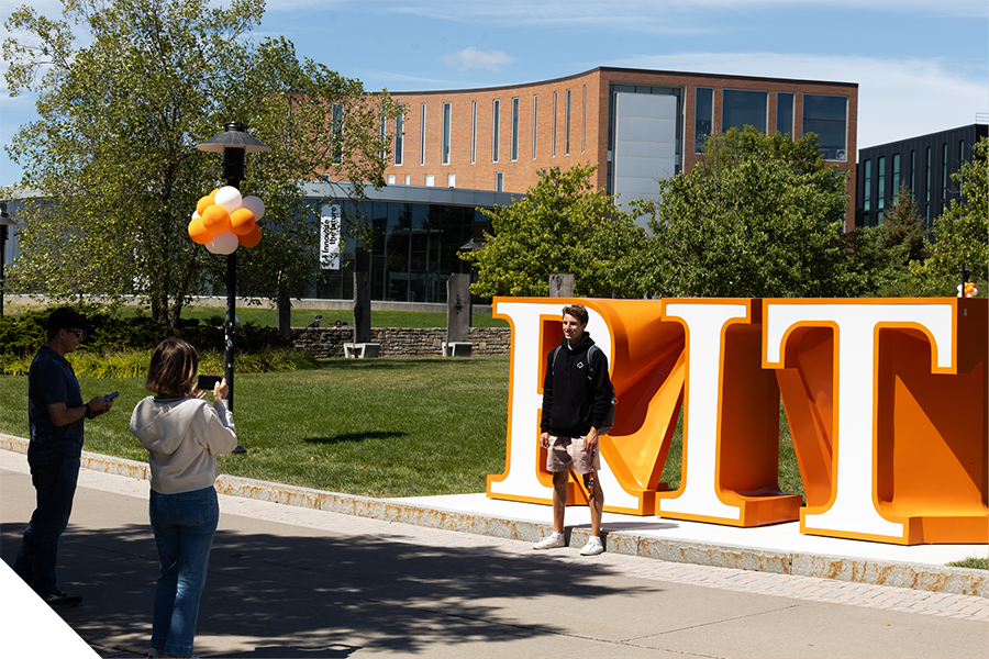 Student stands in front of large orange RIT letters while another person takes a photo on a sunny campus lawn.