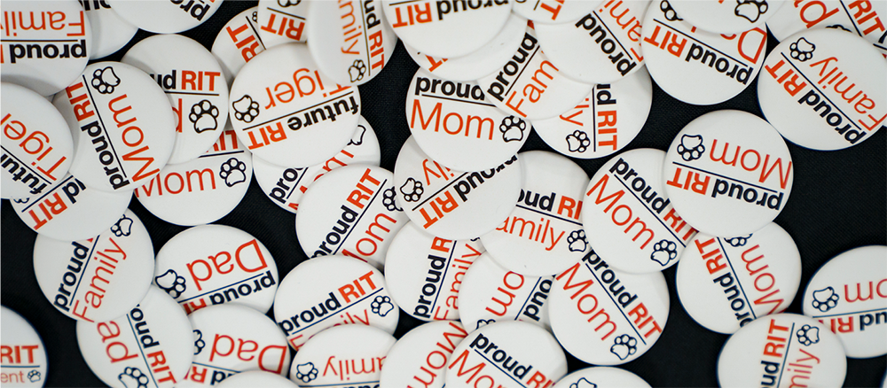 White RIT family buttons scattered across a dark surface, featuring “proud RIT family,” “Mom,” and “Dad” designs with tiger paw icons.