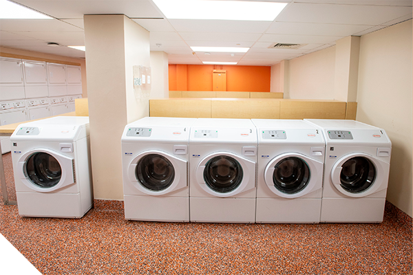 Row of front-loading washing machines in a clean residence hall laundry room, with additional machines along the side wall and an orange accent wall at the back.