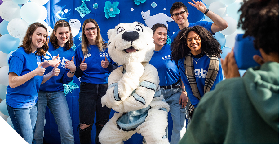 Group of students in blue shirts posing with a white tiger mascot in front of a blue backdrop with winter decorations while someone takes their photo.