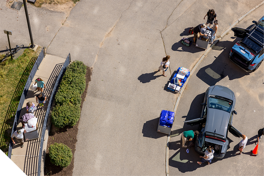Aerial view of move-in day, with multiple vehicles parked along a curb as students and families unload carts and bins filled with belongings. People are moving items between cars and a nearby residence hall entrance on a sunny day.