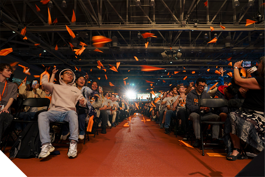 Students toss orange paper airplanes during a large Orientation event in an indoor gathering space.
