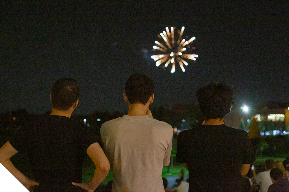 Three students stand outdoors at night watching a burst of fireworks in the sky, with campus lights glowing in the background.