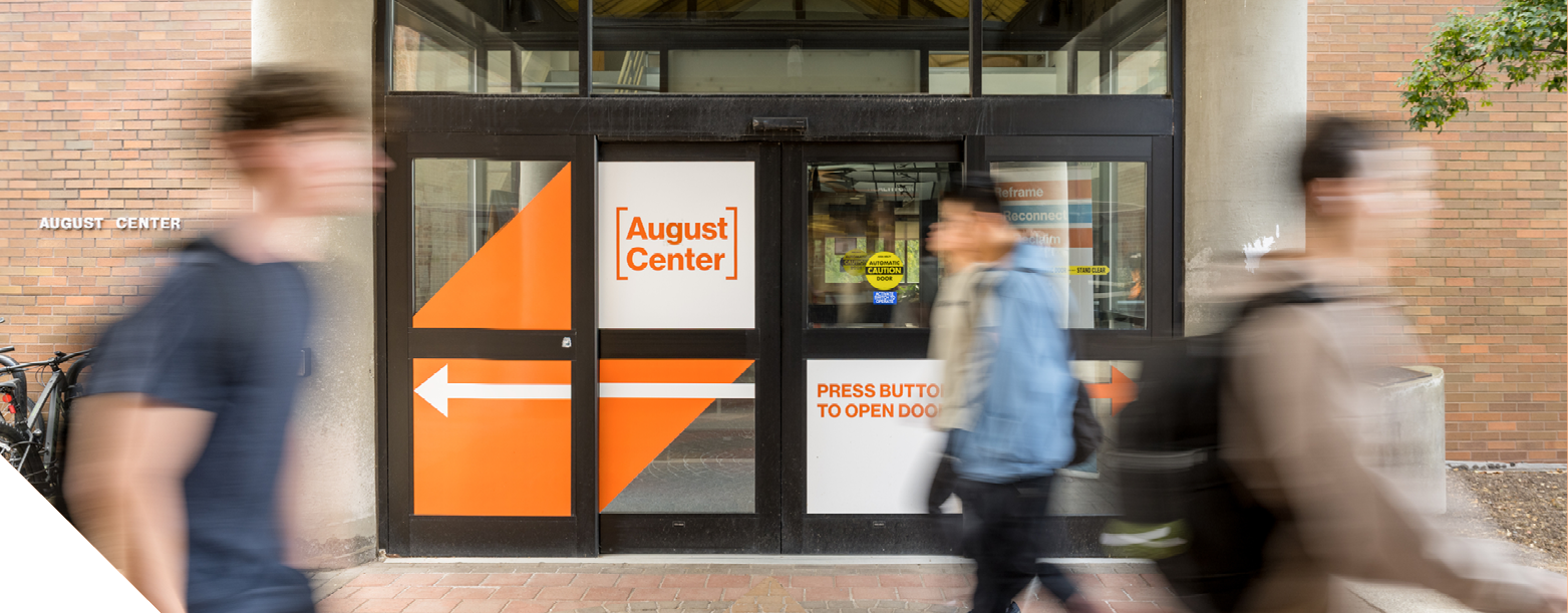 Students walk past the entrance of the August Center, with orange directional graphics and signage on the glass doors.