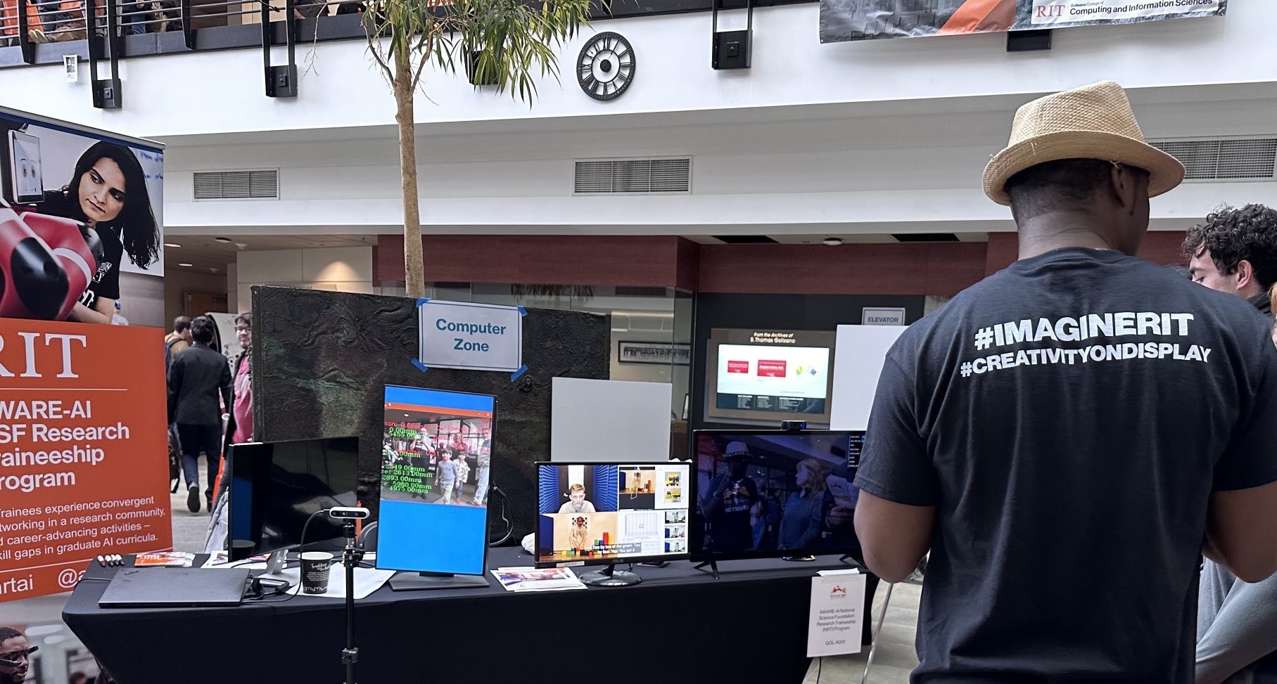 Trainee Justin Namba stands on the right and faces away from the camera to explain about the AWARE-AI NRT to visitors.The large NRT poster sits on the left, with the table with flyers in the middle.