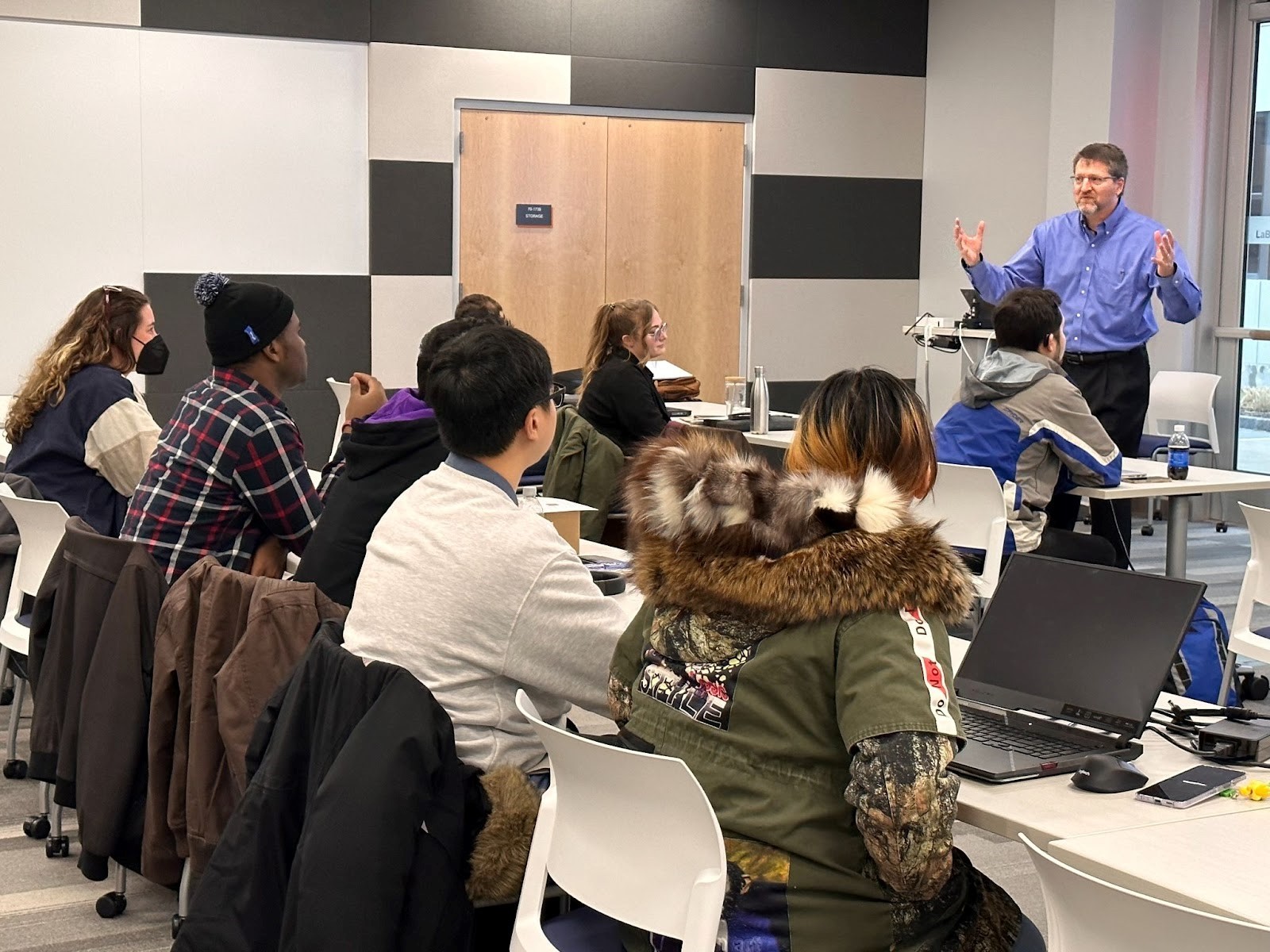 Richard DeMartino, with arms outstretched, faces the students who sit at tables and face away from the camera.
