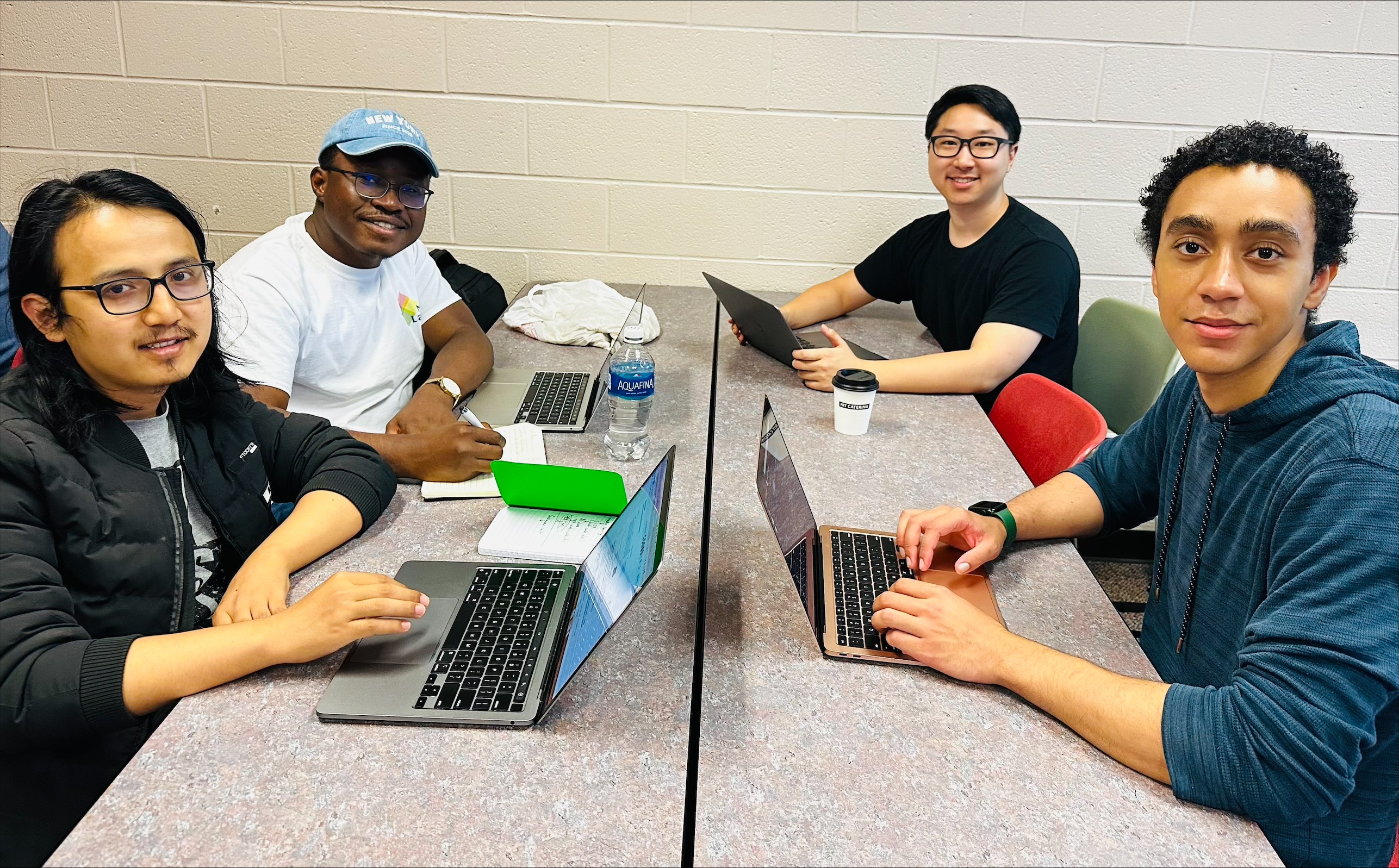 Four students sit around a table with laptops open in front of them, they face the camera and smile.