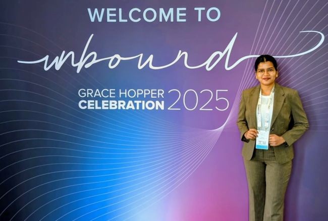 A person stands in front of a “Welcome to Grace Hopper Celebration 2025” backdrop, posing and smiling in professional attire.