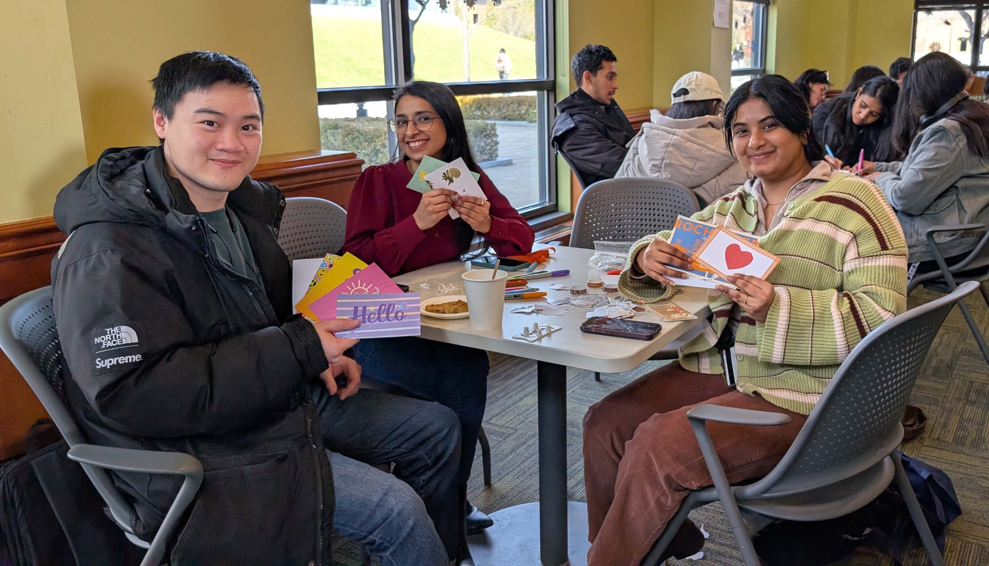 Three students sit at a table during RIT Graduate School’s coffee hour event, smiling and holding postards with art supplies spread out in front of them.