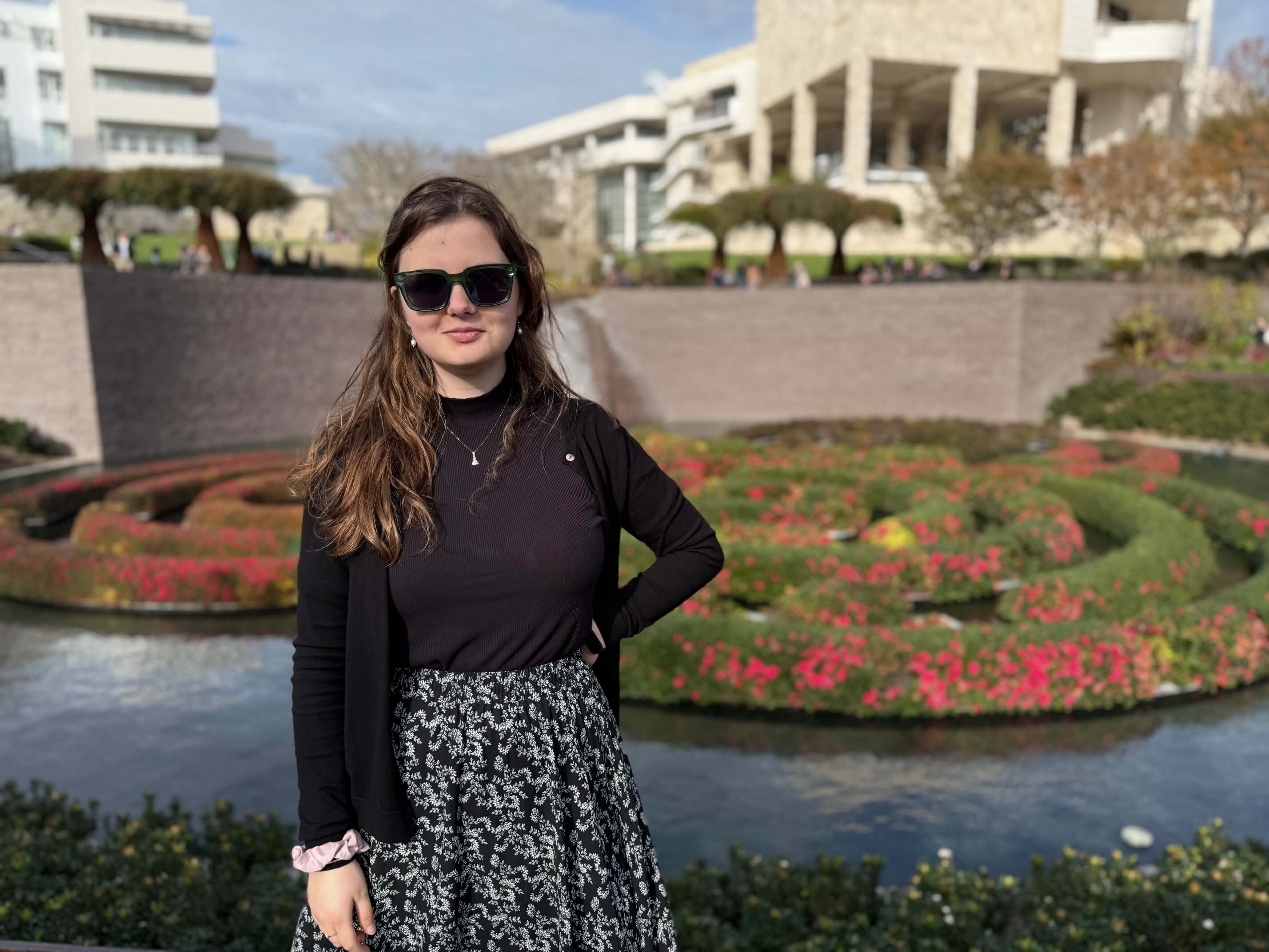 Person wearing sunglasses standing in front of a flower garden with a fountain and buildings in the background.