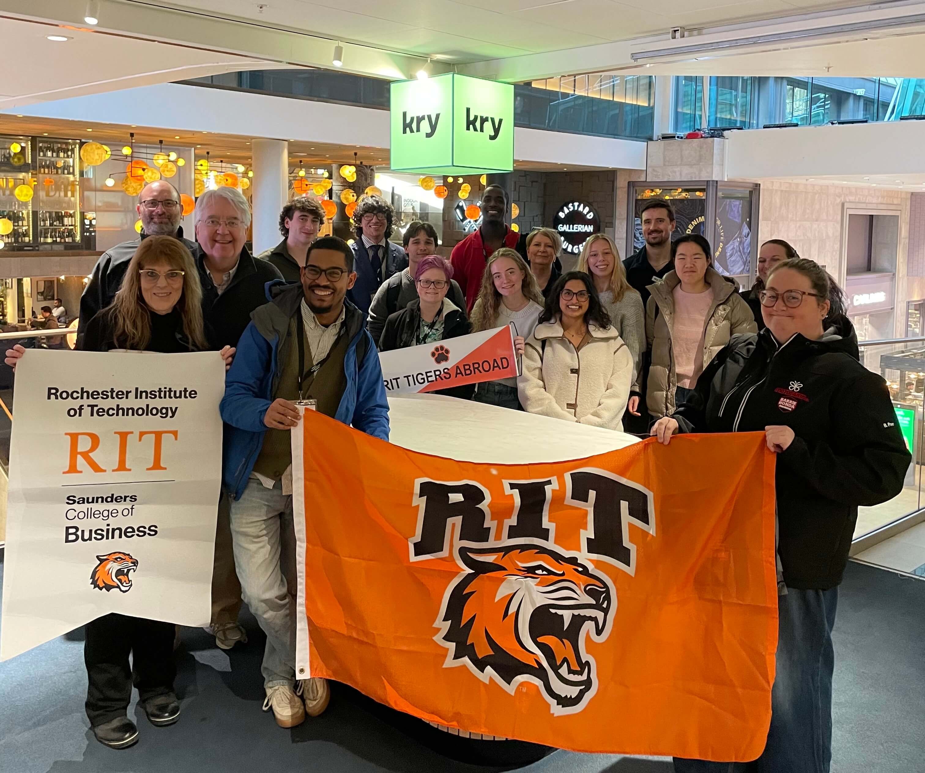 Group of students and staff posing with an orange RIT tiger flag and banners reading “Rochester Institute of Technology” and “RIT Tigers Abroad.”
