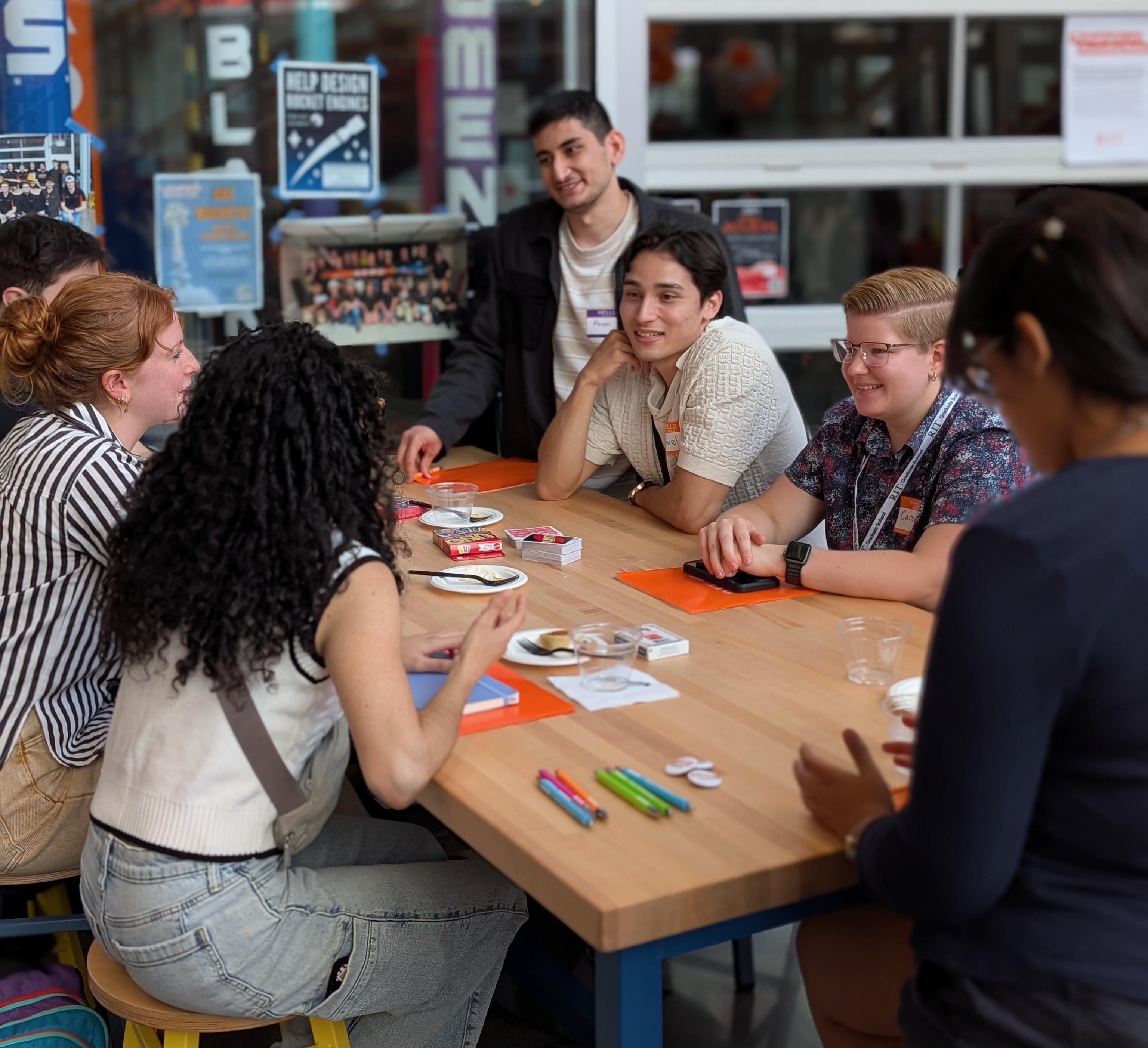 New graduate students sitting on a circular table with one student holding up and showing a gray RIT T-shirt while others smile at the camera